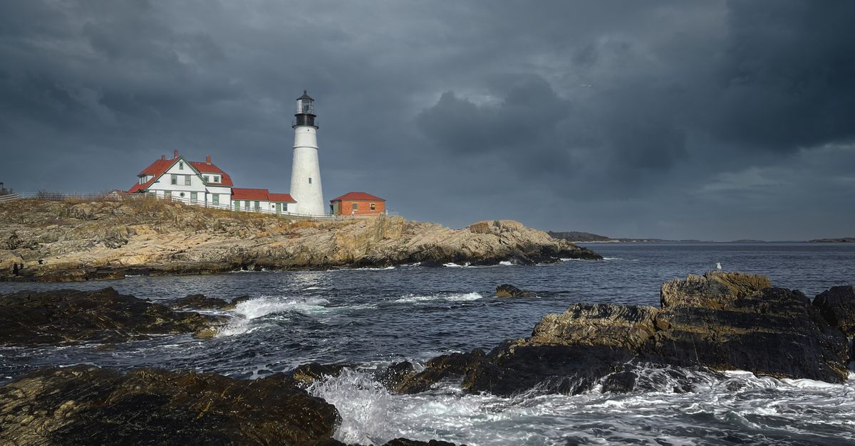 Lighthouse and houses located on shore with rocks washed by foamy waves in gloomy overcast day